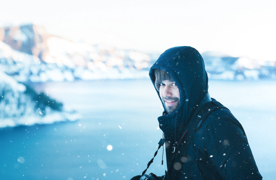 Man in black hooded coat standing in the snow in front of a lake and mountains