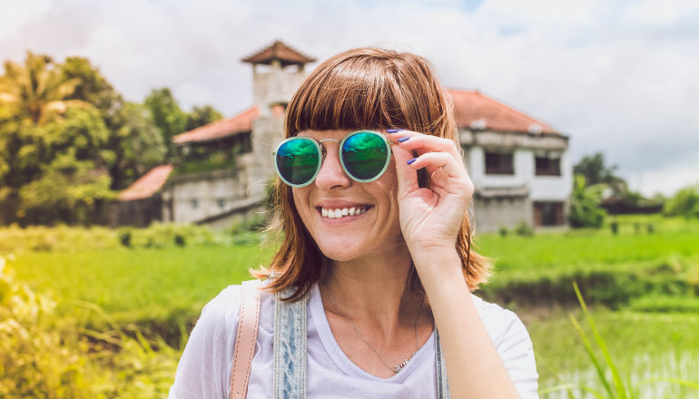 Smiling brunette woman wears blue and green sunglasses outside in a green field by an old building