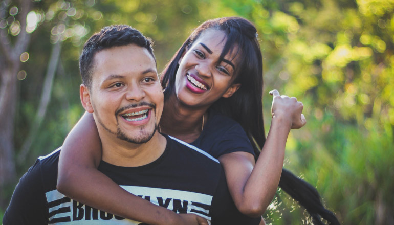 Dark-haired woman embraces a dark-haired man from behind, both smiling and wearing braces