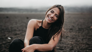 Brunette woman with a black tanktop sits in a dirt field while smiling with a bright white smile thanks to dental veneers