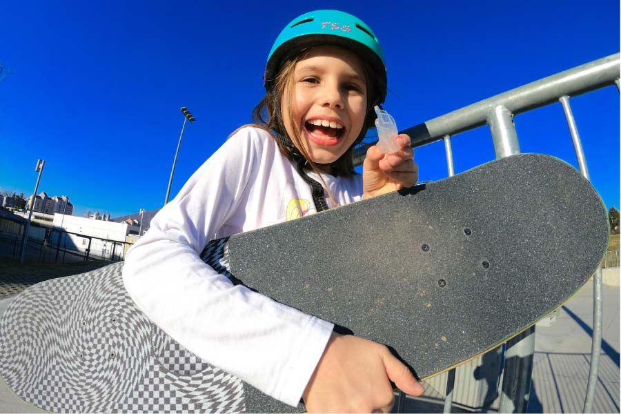 young girl wearing a helmet and holding a skateboard holds up her mouthguard and smiles